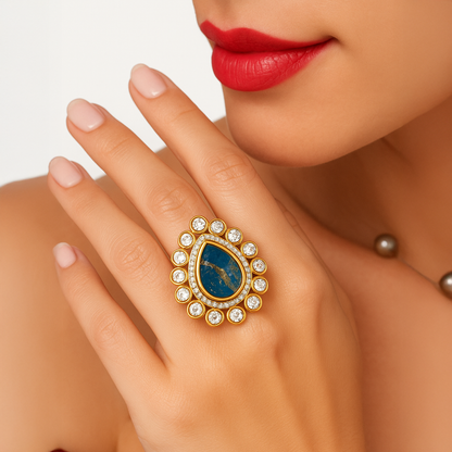 Close-up of a hand wearing a gold ring with a blue stone and diamonds, against a neutral background.