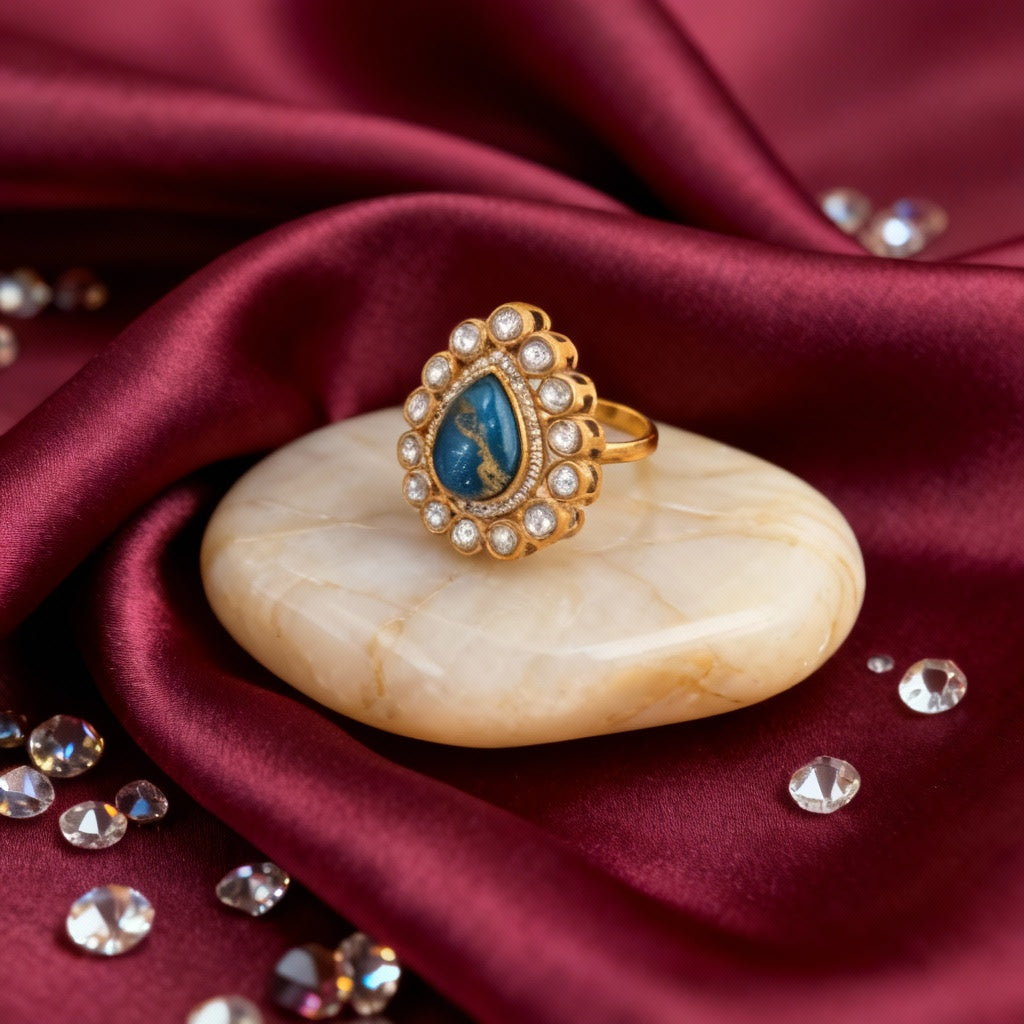 Gold ring with blue gemstone on a beige stone, surrounded by crystals on red fabric