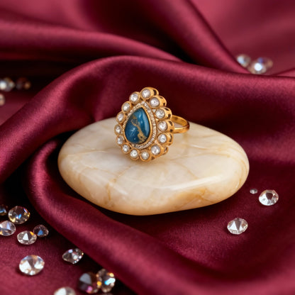 Gold ring with blue gemstone on a beige stone, surrounded by crystals on red fabric