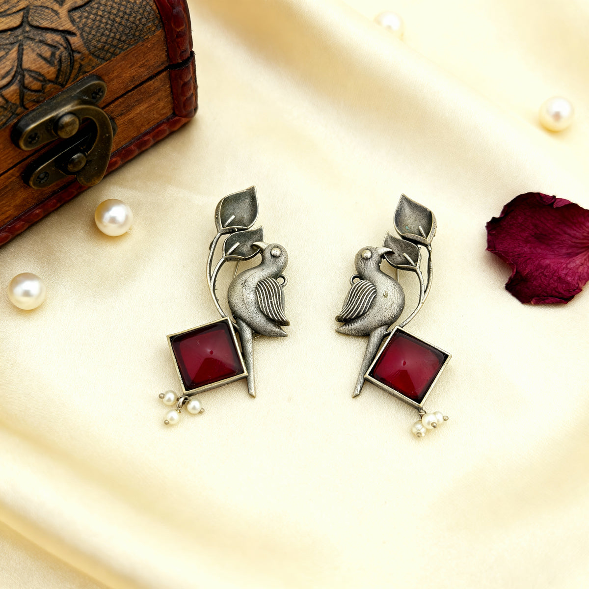 Pair of silver earrings with red stones on a decorative surface with pearls and a wooden box.