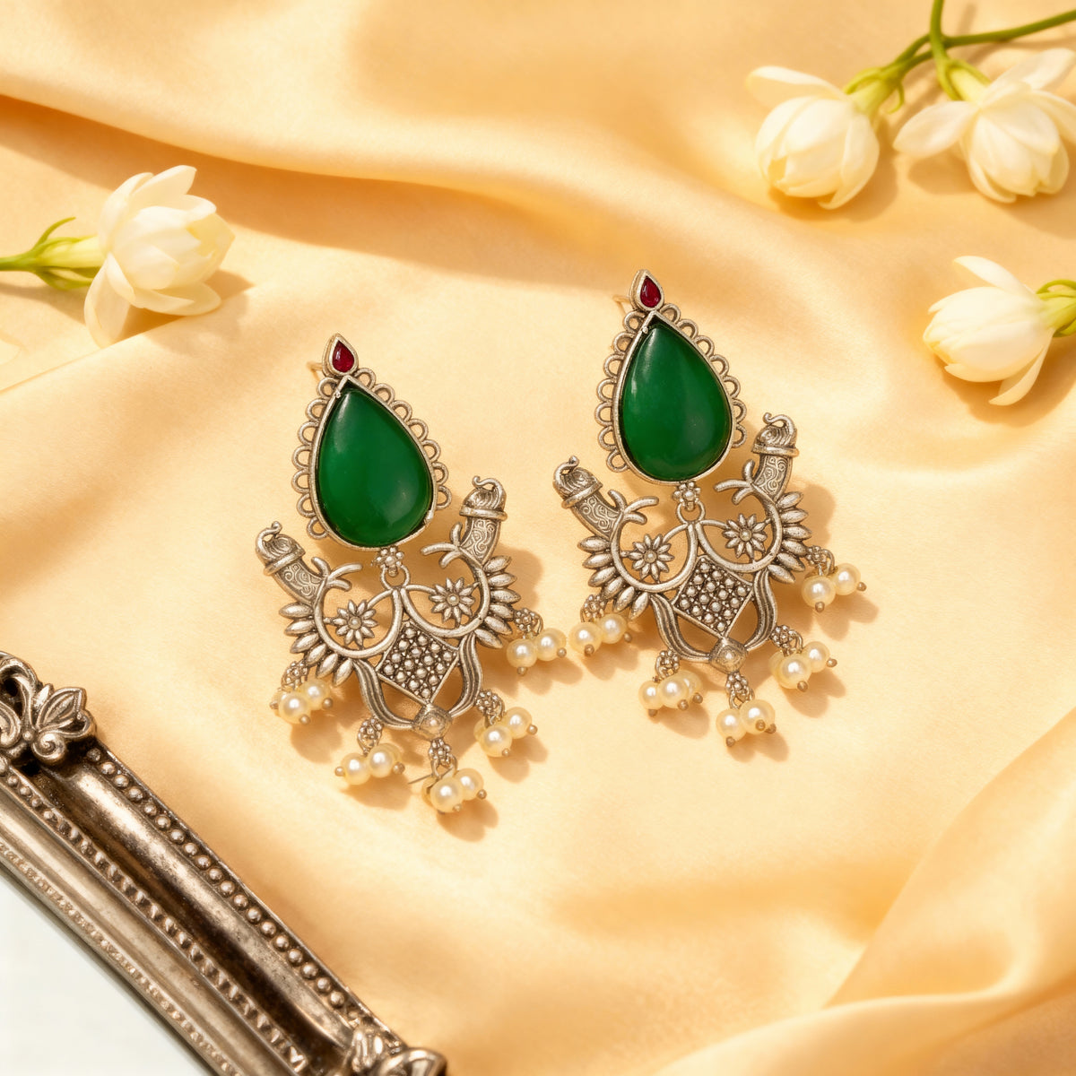 Ornate earrings with green teardrop stones and pearl-like elements on a beige fabric background.
