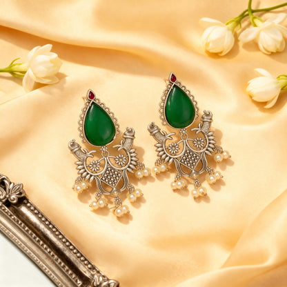 Ornate earrings with green teardrop stones and pearl-like elements on a beige fabric background.