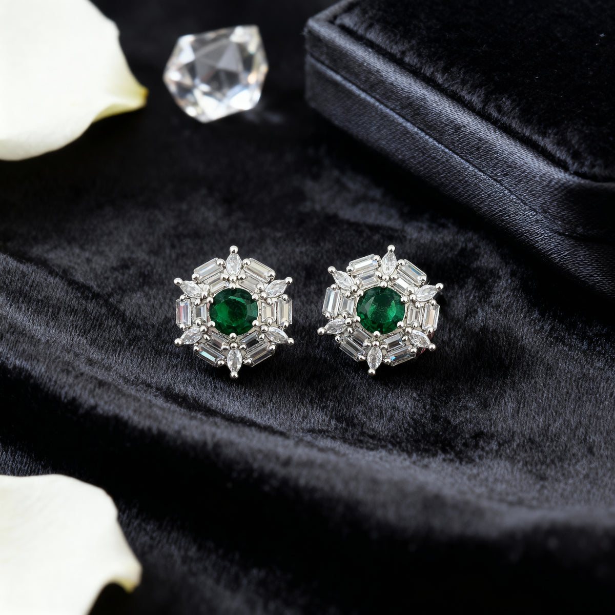 Pair of emerald and diamond stud earrings on a dark fabric background